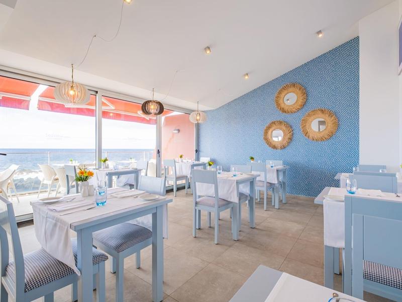 Helles Restaurant mit blau-weißer Einrichtung, Tischen mit weißen Tüchern und großer Fensterfront.