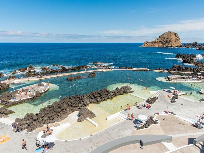 Meerwasserpool mit Felsen, Sonnenschirmen und blauer Meereskulisse am sonnigen Strand.