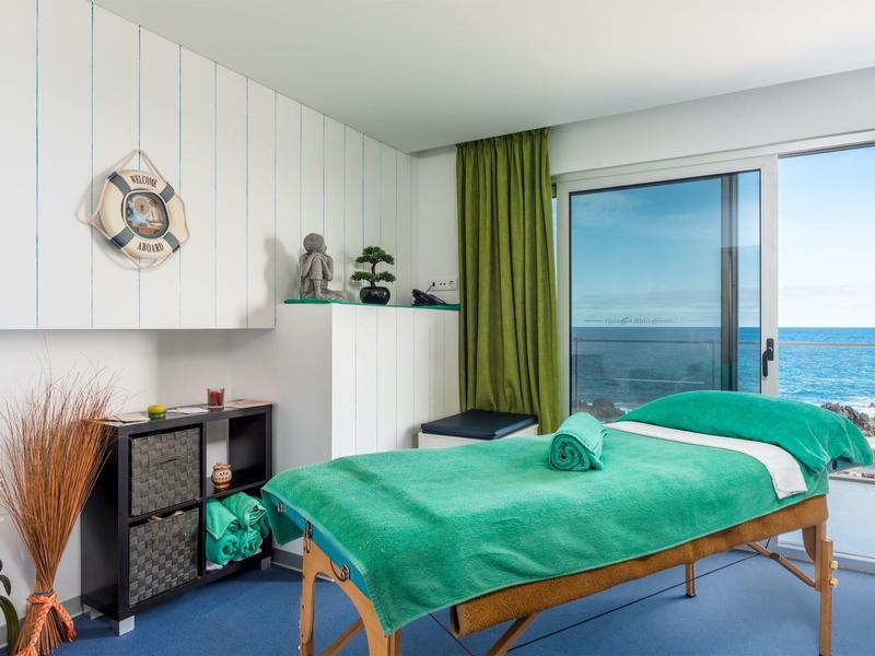 Helles Behandlungszimmer mit Meerblick, grünen Vorhängen und massagetisch mit grünem Bezug.