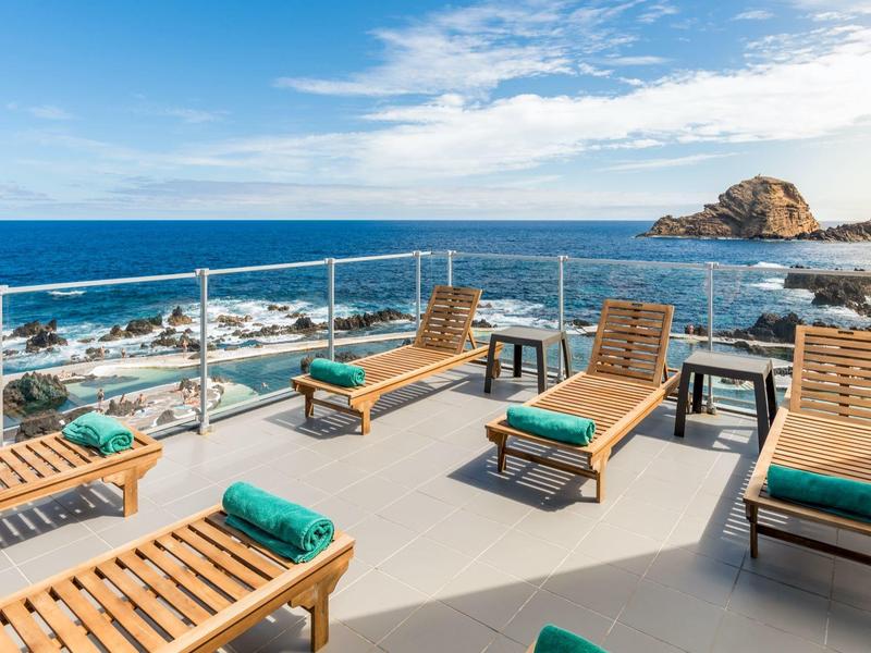 Terrasse mit Holzliegen und grünen Handtüchern, Blick auf das blaue Meer und Felsen bei klarem Himmel.