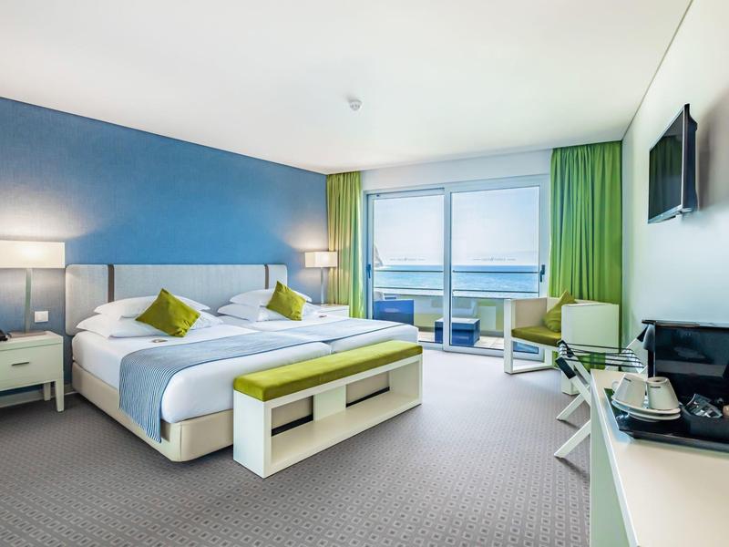 Hell eingerichtetes Hotelzimmer mit Doppelbett, blauer Wand, großem Fenster und grünem Vorhang.