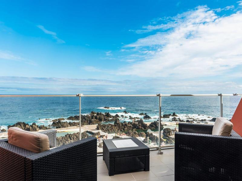 Terrassenblick mit schwarzen Korbsesseln, Glasgeländer, Felsen und blauem Himmel am Meer.