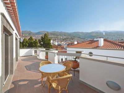 Balcón con sillas y mesa, vista de casas y montañas bajo cielo despejado.