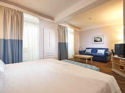 Camera d'hotel luminosa con letto matrimoniale, divano, TV e finestra con tende.