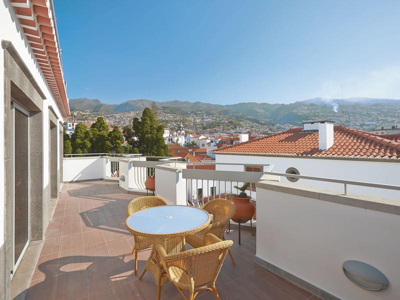 Balcone con tavolo e sedie con vista su montagne e case con tetti rossi.