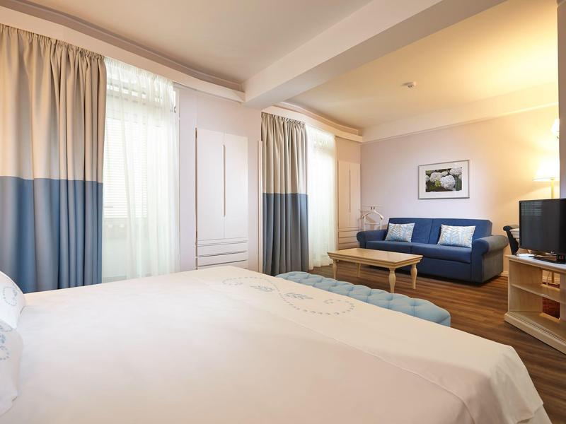 Camera d'hotel luminosa con letto matrimoniale, divano, TV e finestra con tende.