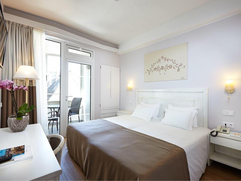 Camera d'hotel luminosa con letto matrimoniale, comodini e balcone con sedie.