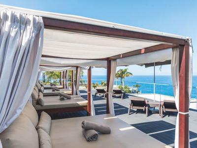 Strandliegen unter Holz-Pavillons mit weißen Vorhängen am Meer bei klarem Himmel.