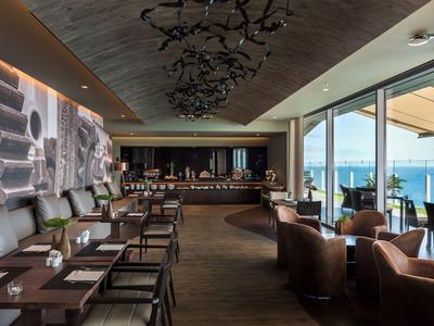 Moderner Restaurantbereich mit braunen Ledersesseln, Holztisch, großer Fensterfront und Meerblick.