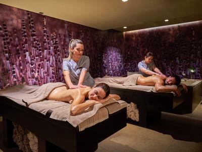 Dunkler Wellnessraum mit violetten Wänden, zwei Liegen und Massagetherapeuten bei Rückenmassage.