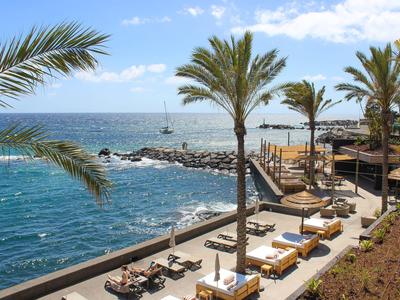 Strandpromenade mit Palmen, Liegestühlen und blauem Meer unter hellem Himmel.