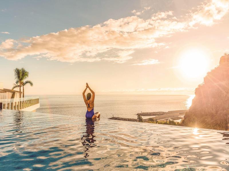 Person macht Yoga in Infinity-Pool bei Sonnenuntergang mit Palmen und Meeresblick.