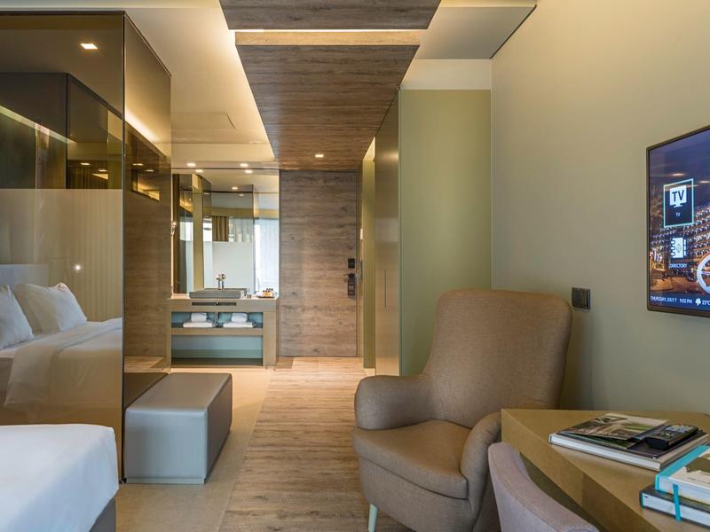 Modernes Hotelzimmer mit Sessel, Bett, TV an der Wand und einem Spiegel im Hintergrund.