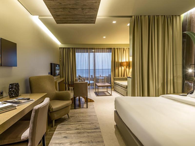 Modernes Hotelzimmer mit großem Bett, Schreibtisch, Sessel und Balkon mit Meerblick bei Dämmerung.