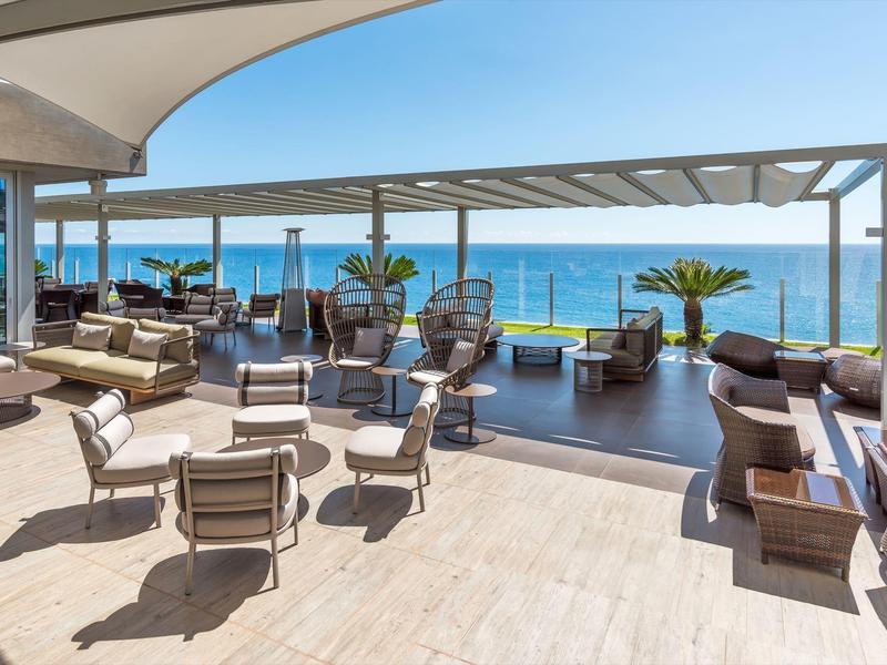 Offener Terrassenbereich mit modernen Sitzmöbeln, Palmen und Meerblick bei klarem Himmel.