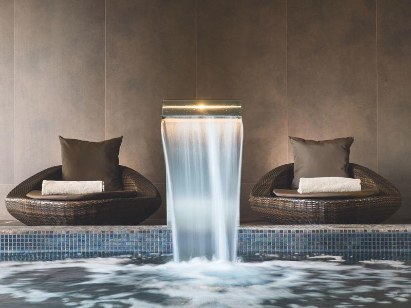 Moderner Innenpool mit beleuchtetem Wasserfall und zwei runden Sesseln mit Kissen an brauner Wand.