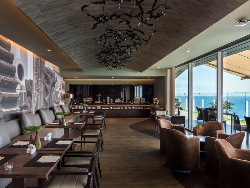 Moderner Restaurantbereich mit braunen Ledersesseln, Holztisch, großer Fensterfront und Meerblick.