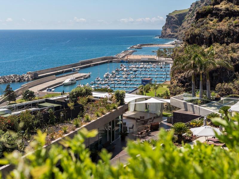 Küstenlandschaft mit Hafen, Booten, grünen Pflanzen und Klippen unter blauem Himmel.