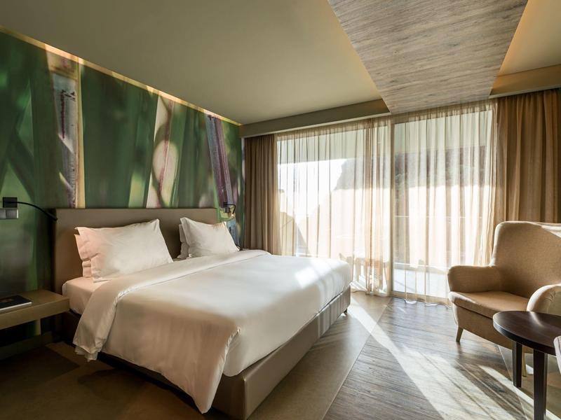 Helles Schlafzimmer mit großem Bett, grünem Wanddekor, Sessel und bodentiefen Fenstern mit Vorhängen.