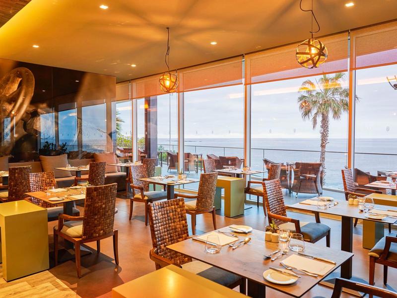 Helles Restaurant mit Holzstühlen, großen Fenstern und Meerblick bei Sonnenuntergang.