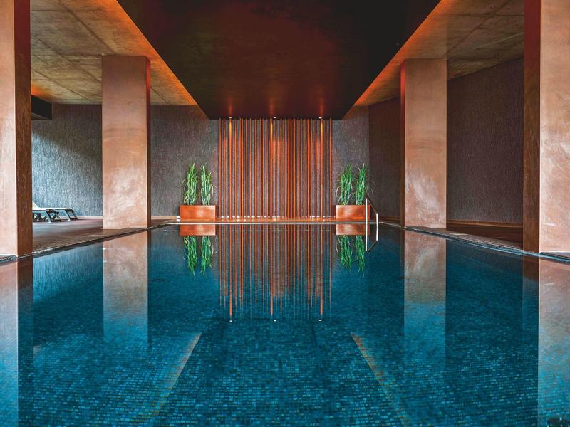 Moderner Innenpool mit blauem Wasser, warmen Braun- und Orangetönen, Säulen und Pflanzen am Beckenrand.