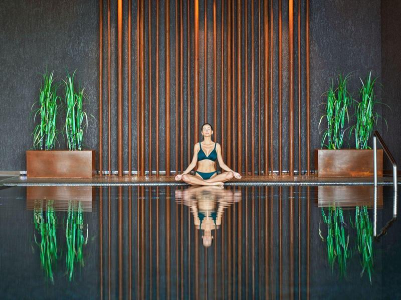 Frau mit Sportkleidung sitzt überkreuzt am Rand eines stillen Pools vor holzverkleinter Wand und Pflanzen.