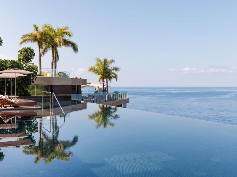 Infinity-Pool mit Palmen, Liegestühlen und Blick auf ruhiges, blaues Meer unter klarem Himmel.
