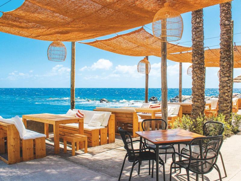 Strandterrasse mit Holz- und Metallmöbeln, orange Segeltuchdach und Blick auf das blaue Meer.