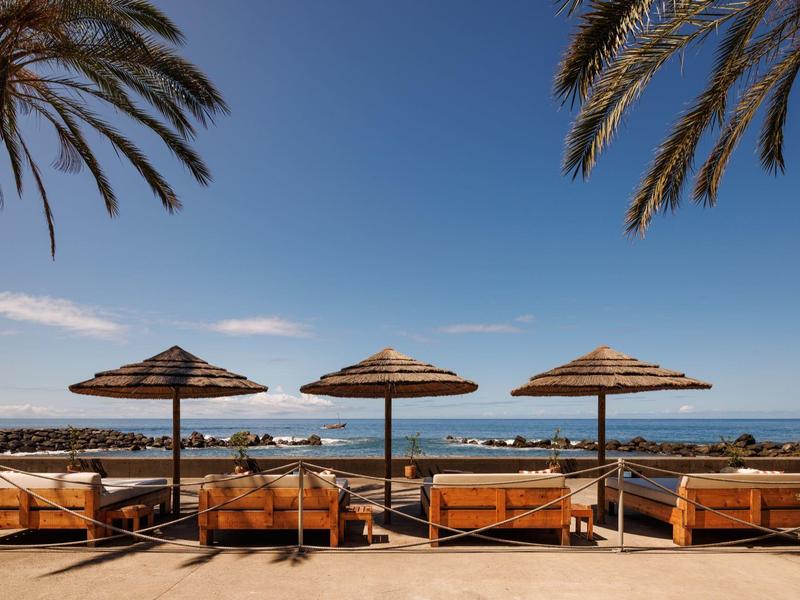 Strandterrasse mit vier Sonnenschirmen, Holzliegen und Blick aufs blaue Meer unter klarem Himmel.