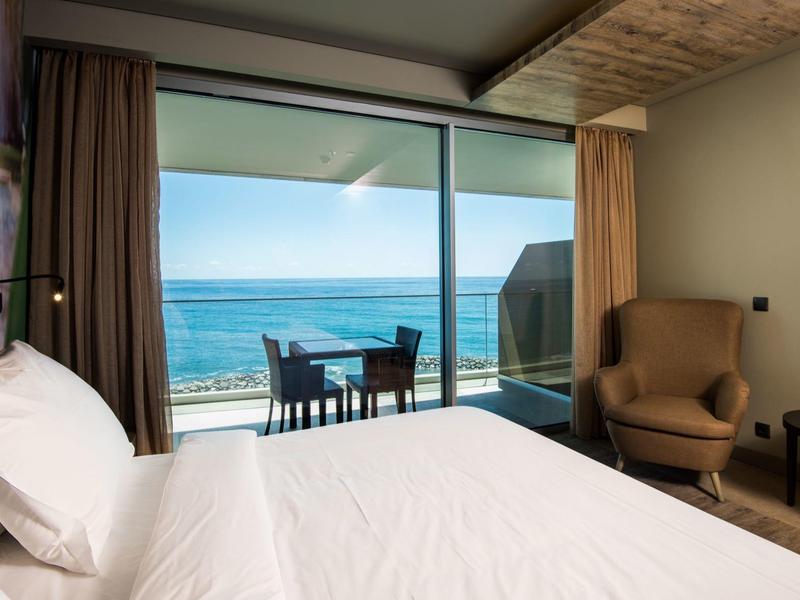 Helles Schlafzimmer mit großem Bett, Sessel, Tisch und Meerblick durch raumhohe Fenster und Terrasse.