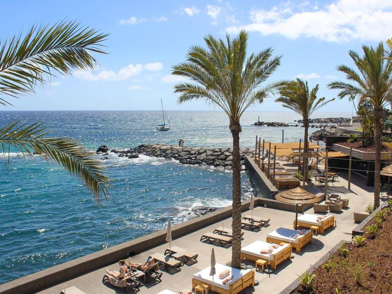 Strandpromenade mit Palmen, Liegestühlen und blauem Meer unter hellem Himmel.