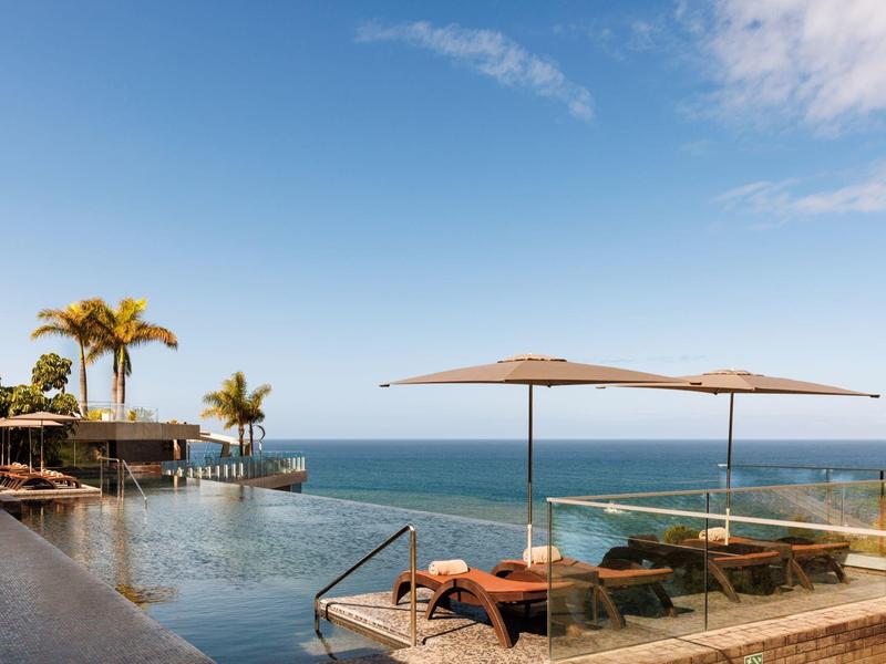 Infinity-Pool mit Blick aufs Meer, Sonnenliegen und Sonnenschirme unter klarem Himmel.