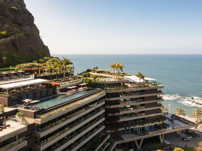 Moderner Hotelkomplex mit mehreren Terrassen, Palmen und Meeresblick an felsiger Küste bei klarem Himmel.