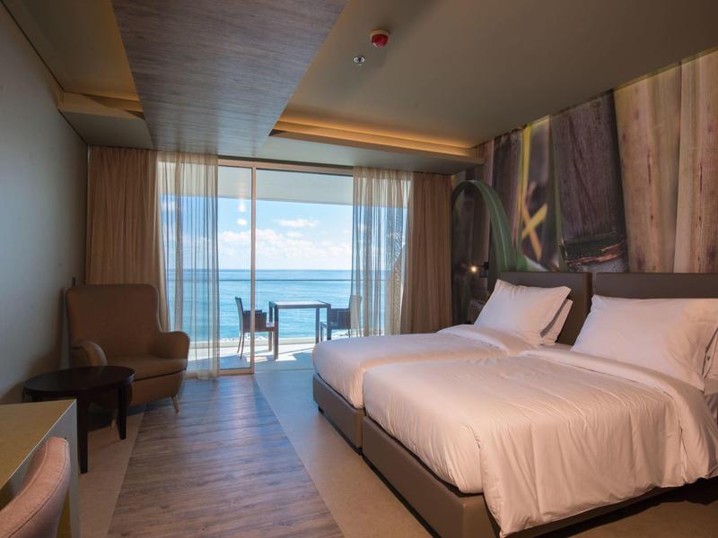 Modernes Hotelzimmer mit Doppelbett, Lehnsessel und Balkon mit Meerblick bei Tageslicht.