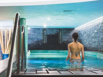 Woman in bikini stands in modern spa pool with stone walls and subdued lighting.