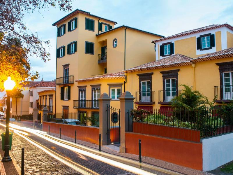 Straße mit Kopfsteinpflaster und gelbem Haus mit grünen Fensterläden bei Abendlicht.