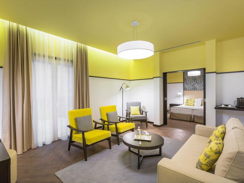 Helles Wohnzimmer mit gelben Akzenten, bequemen Sesseln, Sofa und rundem Couchtisch vor Vorhängen.