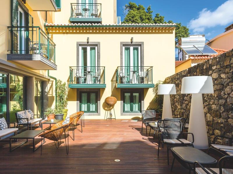 Sonnige Terrasse mit Holzboden, Sitzgelegenheiten und gelbem Gebäude im Hintergrund.