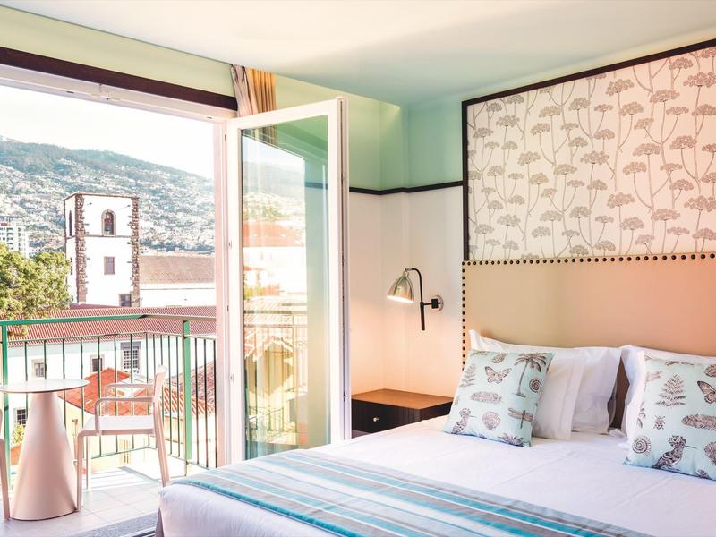 Helles Schlafzimmer mit Doppelbett und offenem Balkon, Bergblick und hellgrünen Wänden.