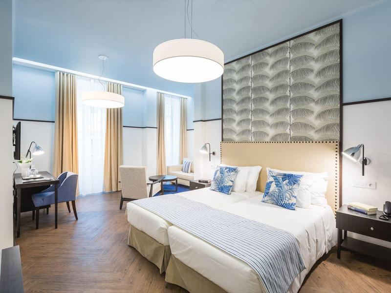 Modernes Hotelzimmer mit blau-weißer Farbgebung, Doppelbett, Schreibtisch und großen Fenstern.