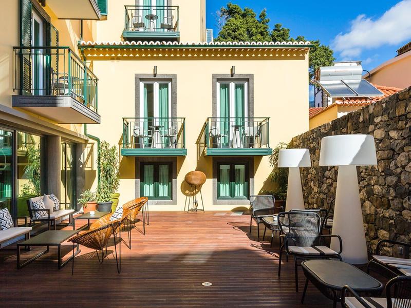 Sonnige Terrasse mit Holzboden, Sitzmöbeln, weißen Lampen und gelbem Haus im Hintergrund.