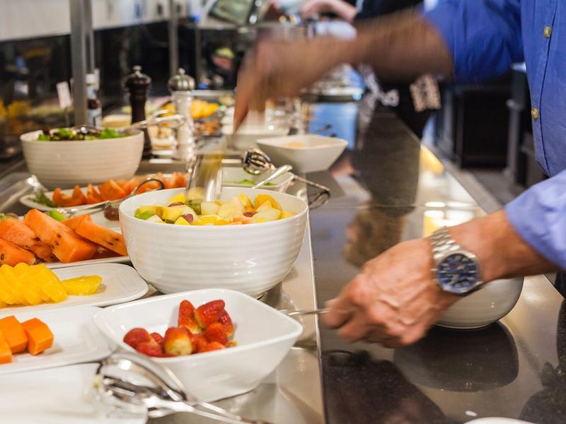 Buffet mit weißen Schalen voll Gemüse und Salat, Person in blauem Hemd greift mit Hand zu.