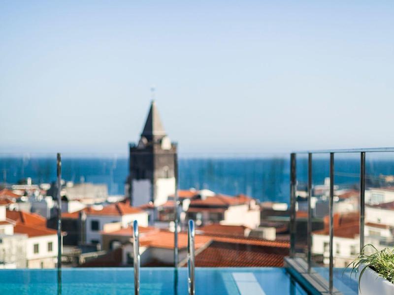 Stadt mit roten Dächern, Kirchturm und dahinter blaues Meer unter klarem Himmel.