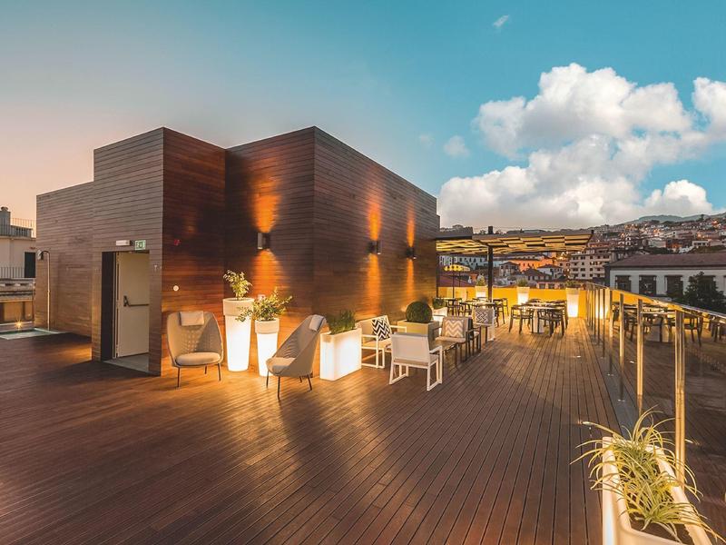 Moderne Dachterrasse mit Holzboden, Sitzgruppen, Pflanzen und Beleuchtung im Abendlicht.