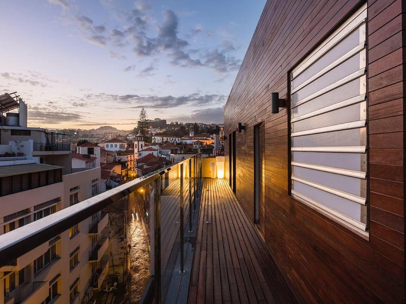 Balkon mit Holzverkleidung und Glasgeländer bei Sonnenuntergang über Stadtblick.