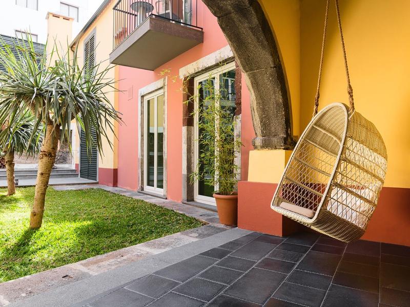 Moderner Außenbereich mit gelber Wand, Hängekorb, Pflanze und kleinen Bäumen auf grünem Rasen.