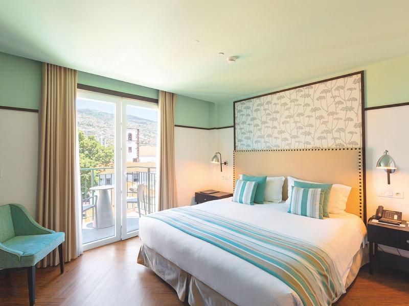 Helles Schlafzimmer mit Doppelbett, türkis-weißer Bettwäsche, Sessel und Balkon mit Glas­tür.