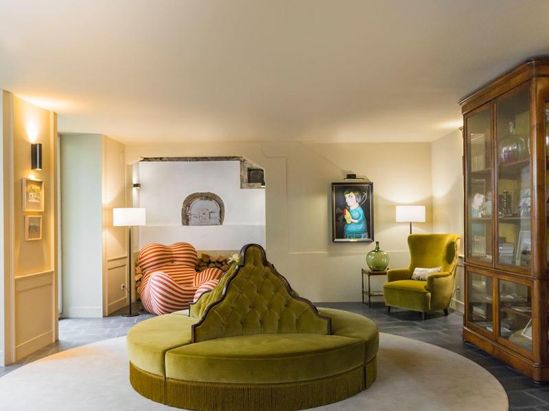 Gemütliches Wohnzimmer mit runder grüner Couch, Sessel, Kamin und Holzvitrine, helle Wände, warmes Licht.