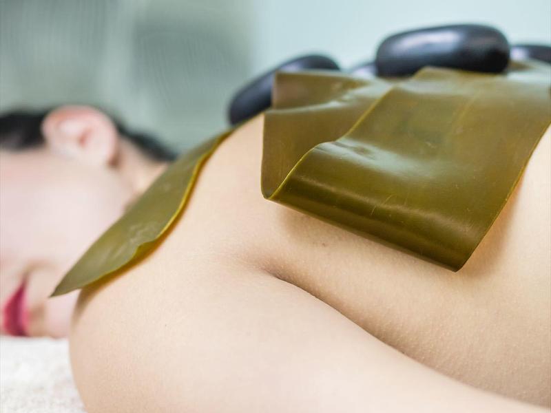 Frau entspannt auf Bauch, warme Steine und Algenblätter liegen auf ihrem Rücken, Wellness-Atmosphäre.