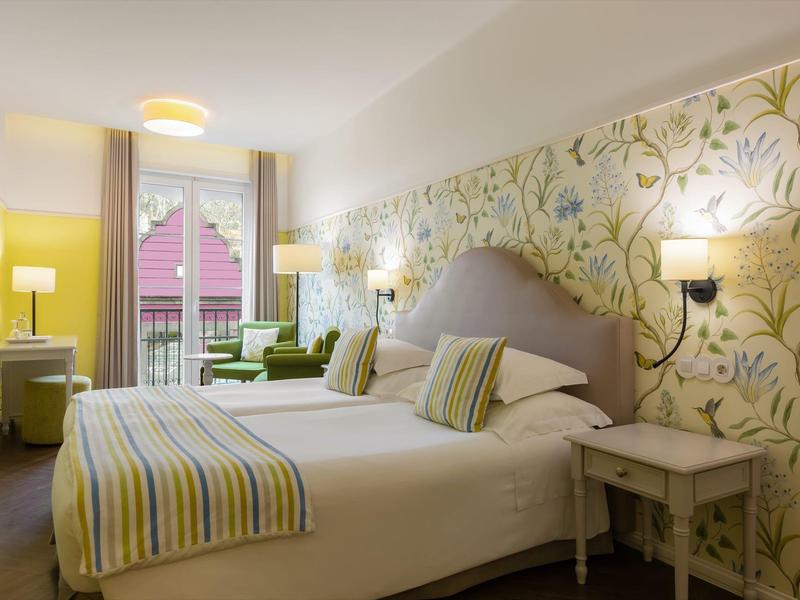 Helles Schlafzimmer mit großem Bett, gestreiften Textilien und floralem Tapetenmuster an der Wand.